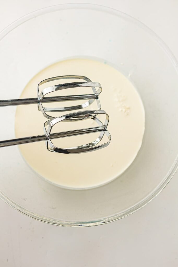 Metal hand mixer beaters resting inside a large glass mixing bowl filled with cold heavy cream before whipping homemade frosting for patriotic red, white, and blue cupcakes. The cream is smooth and liquid at this stage before being beaten into soft, fluffy peaks.