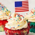 Close-up of a patriotic red, white, and blue cupcake topped with a swirl of fluffy whipped cream frosting, red white and blue sprinkles, and a small American flag pick. The cupcake features visible layers of bright blue cake on the bottom with red marbled cake on top, while additional frosted cupcakes appear softly blurred in the background. This image shows the finished decorated cupcakes ready for serving at a 4th of July celebration.