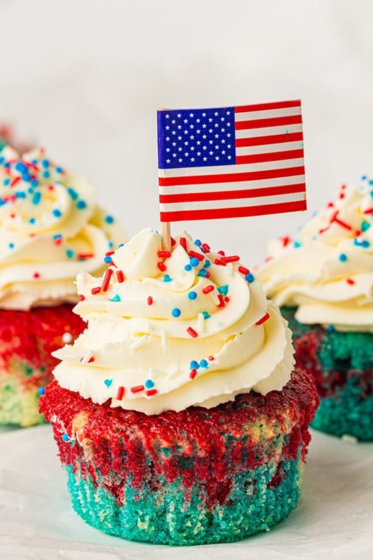 Close-up of a patriotic red, white, and blue cupcake topped with a swirl of fluffy whipped cream frosting, red white and blue sprinkles, and a small American flag pick. The cupcake features visible layers of bright blue cake on the bottom with red marbled cake on top, while additional frosted cupcakes appear softly blurred in the background. This image shows the finished decorated cupcakes ready for serving at a 4th of July celebration.