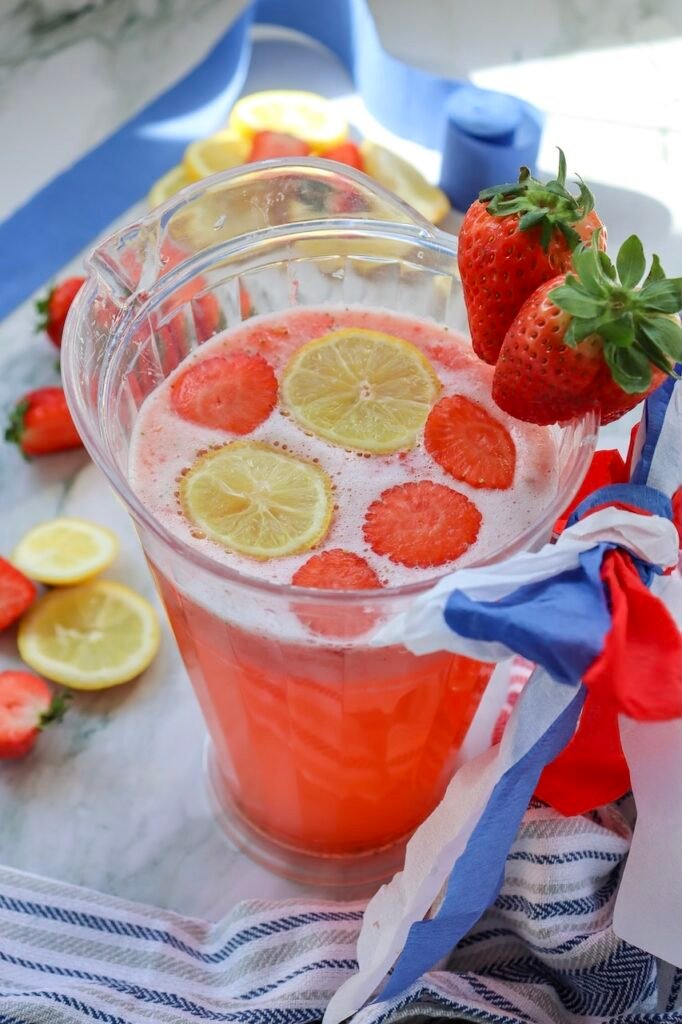 Clear pitcher of patriotic strawberry lemonade topped with floating lemon slices and strawberry slices, styled with fresh strawberries and red, white, and blue ribbon decorations for a festive summer party drink.