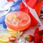 Glass of patriotic strawberry lemonade with ice and lemon slices served beside fresh strawberries and a pitcher decorated with red, white, and blue ribbon for a festive 4th of July summer drink.