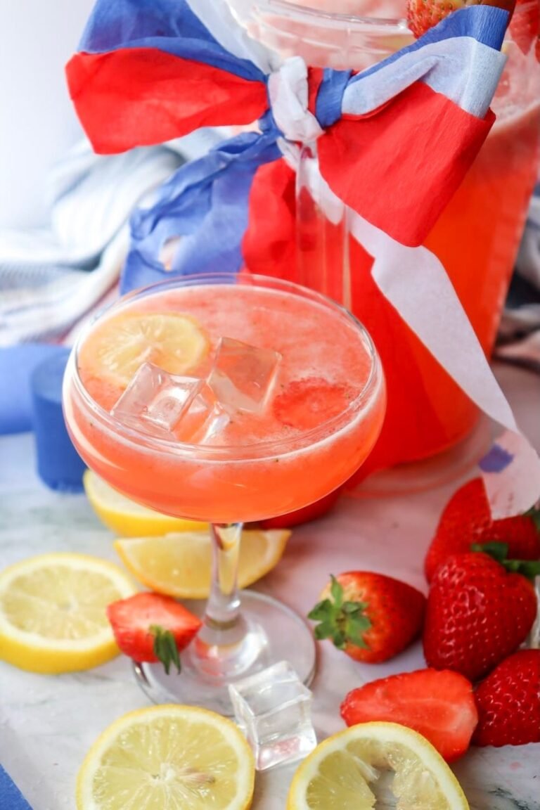 Glass of patriotic strawberry lemonade with ice and lemon slices served beside fresh strawberries and a pitcher decorated with red, white, and blue ribbon for a festive 4th of July summer drink.
