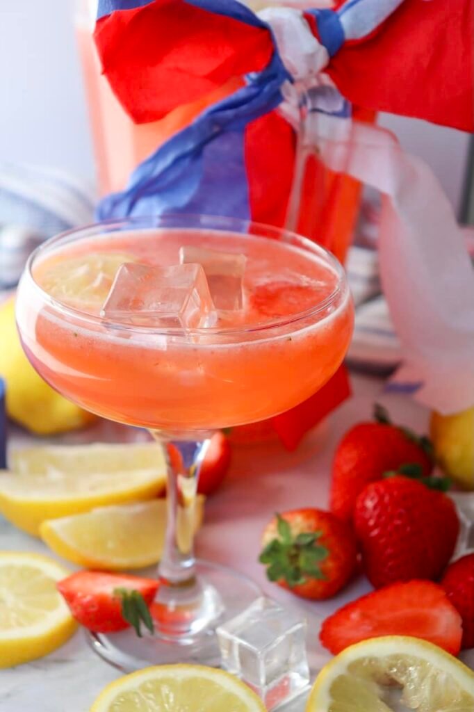 Glass of patriotic strawberry lemonade served over ice with lemon slices and fresh strawberries nearby, styled with red, white, and blue patriotic ribbon decorations for a festive summer party drink.