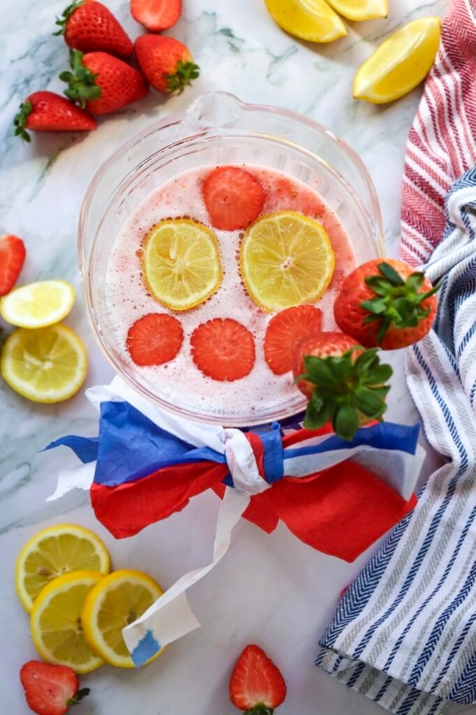 Overhead view of patriotic strawberry lemonade in a clear pitcher topped with floating lemon slices and strawberry slices, surrounded by fresh strawberries, lemon wedges, striped kitchen towels, and red, white, and blue patriotic decorations on a marble countertop.