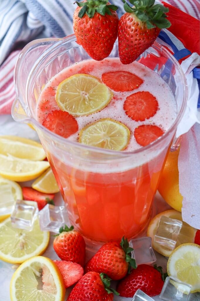 Pitcher of patriotic strawberry lemonade filled with fresh lemon slices and strawberry slices, surrounded by whole strawberries, ice cubes, and patriotic red, white, and blue decorations on a summer party table.