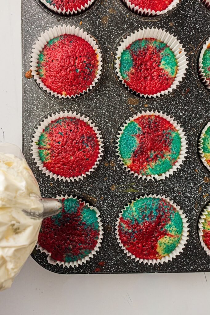 Freshly baked patriotic cupcakes in white paper liners sitting in a dark speckled muffin pan while whipped cream frosting is being piped onto the tops using a pastry bag fitted with a large star tip. The cupcakes have vibrant red and blue swirled tops with a marbled tie-dye effect, showing the decorating step before adding festive red, white, and blue sprinkles.