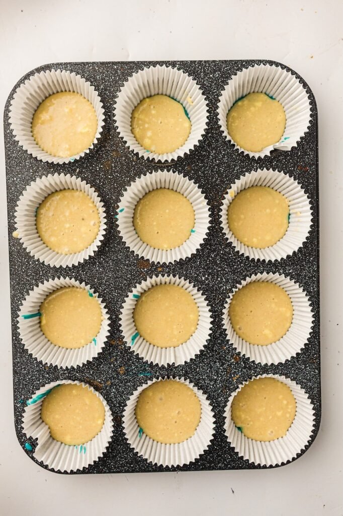Plain vanilla cupcake batter spooned over the blue batter layer inside white cupcake liners arranged in a dark speckled muffin pan. The smooth pale batter creates the middle layer for homemade red, white, and blue 4th of July cupcakes before the final red batter is added on top.