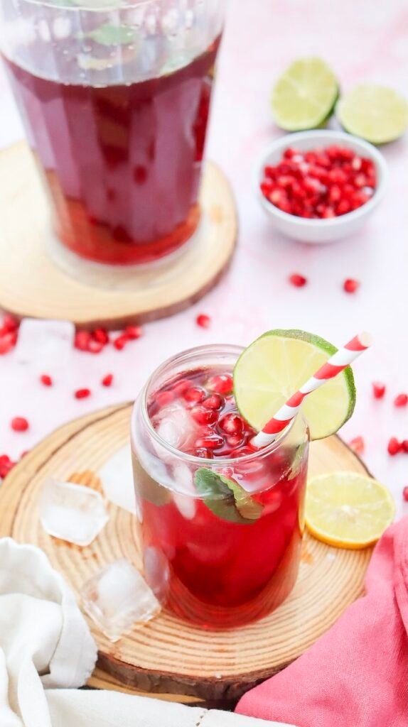 Sparkling pomegranate fizz mocktail served over ice with fresh mint, lime slices, and pomegranate seeds beside a pitcher of the drink on a wooden serving board.