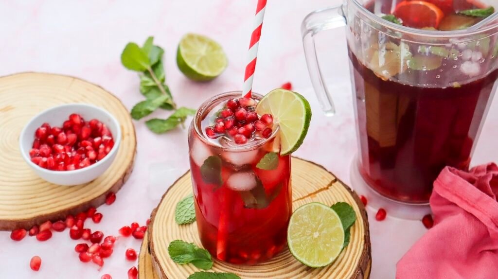 Glass of sparkling pomegranate fizz mocktail garnished with fresh pomegranate seeds, mint, lime, and a striped straw beside a pitcher of the drink on a light pink background.
