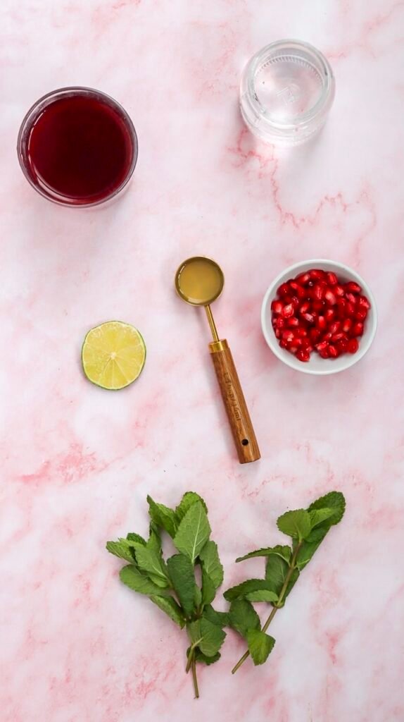 Ingredients arranged on a pink marble surface for a pomegranate fizz mocktail, including pomegranate juice, soda water, fresh lime, pomegranate seeds, mint leaves, and lime juice.