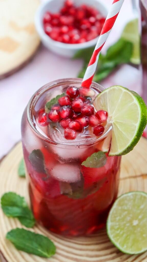 Close-up of a sparkling pomegranate fizz mocktail served over ice with fresh mint leaves, bright pomegranate seeds, and a lime slice garnish in a clear glass with a red striped straw on a wooden serving board.