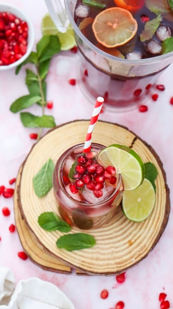 Sparkling pomegranate fizz mocktail served over ice with fresh mint, lime slices, and pomegranate seeds beside a pitcher of the drink on a wooden serving board.
