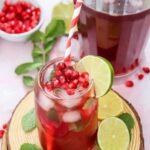 Glass of sparkling pomegranate fizz mocktail filled with ice, fresh mint, lime slices, and pomegranate seeds beside a pitcher of the drink.