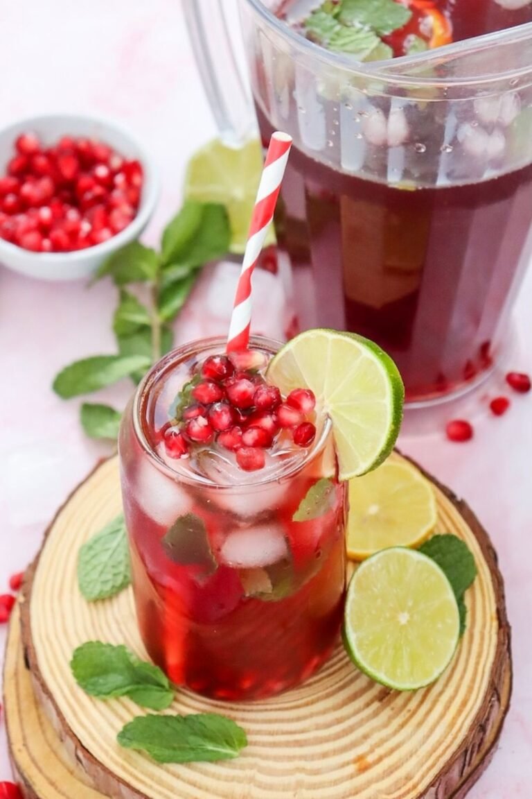 Glass of sparkling pomegranate fizz mocktail filled with ice, fresh mint, lime slices, and pomegranate seeds beside a pitcher of the drink.