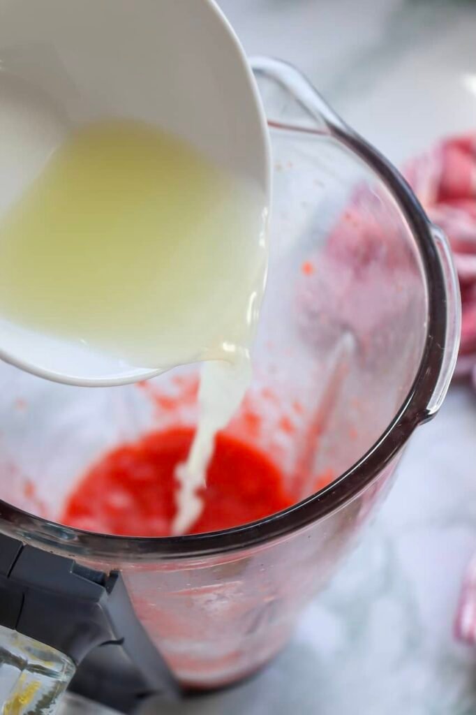 fresh lemon juice being poured from a white bowl into blended puree inside a blender while making homemade drink, with soft pink strawberry tones visible throughout the mixture and blurred fresh strawberries in the background.