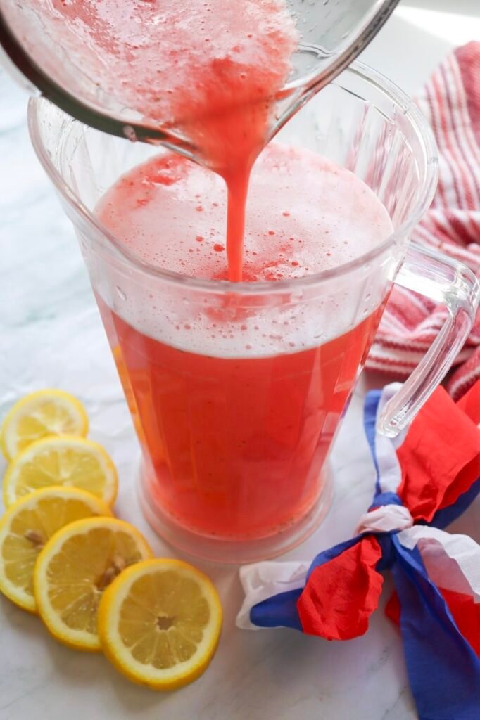 Fresh drink being poured from a blender into a clear pitcher, creating a bright pink-red summer drink with sliced lemons and patriotic red, white, and blue decorations nearby on a marble countertop.