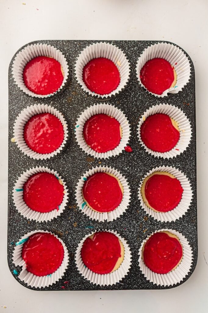 Bright red cupcake batter spooned over a plain vanilla batter layer inside white cupcake liners arranged in a dark speckled muffin pan. Small edges of the pale vanilla batter and blue bottom layer are still visible around some liners, showing the layered red, white, and blue cupcake batter before baking.