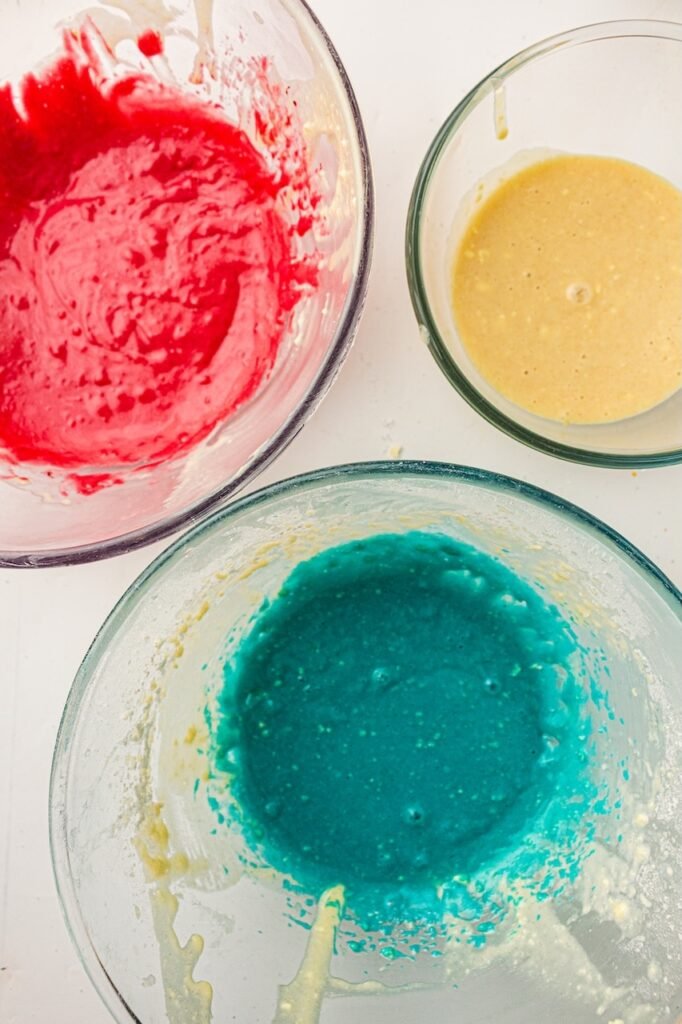 Three clear glass bowls filled with cupcake batter on a white surface, including bright red batter, blue batter, and plain vanilla batter prepared for layered 4th of July cupcakes. The colored batters have a smooth texture with vibrant patriotic colors ready to be spooned into cupcake liners.