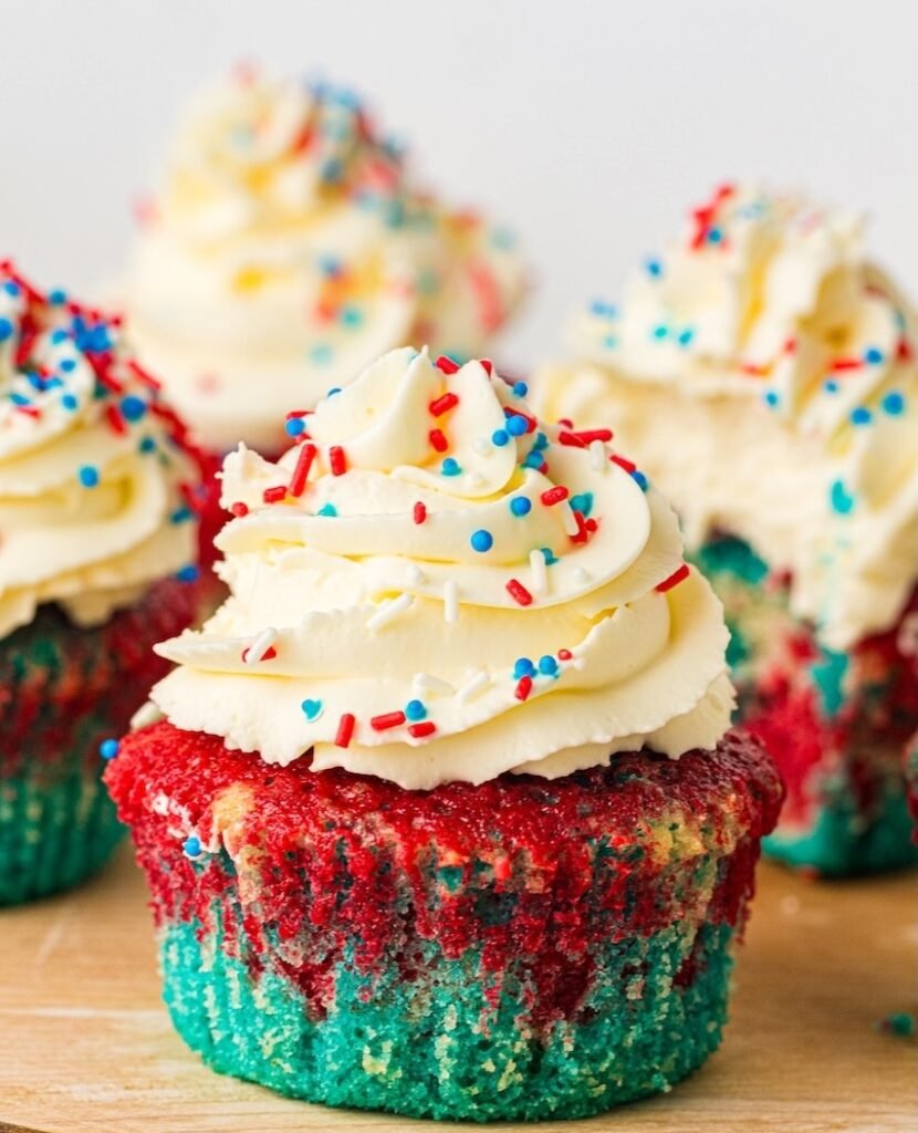 Close-up hero image of a patriotic red, white, and blue cupcake topped with swirled whipped cream frosting and red, white, and blue sprinkles on a wooden serving board. The cupcake liner shows vibrant blue cake on the bottom and bright red cake on top with a marbled vanilla center, while several frosted cupcakes blur softly in the background. This finished 4th of July cupcake recipe is decorated for summer parties, Memorial Day, and Independence Day celebrations.