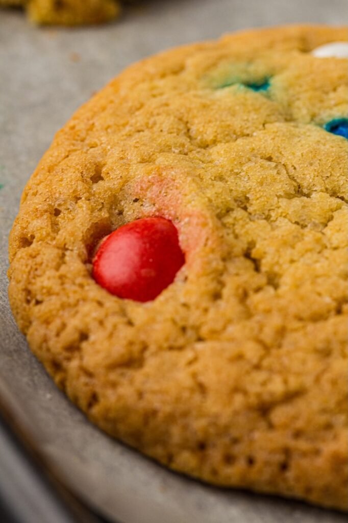 Close-up of a baked cookie on a baking tray with golden brown edges, soft chewy texture, and red, white, and blue candies baked into the surface. The homemade patriotic cookie has a thick bakery-style appearance with slightly crisp edges and a soft center.