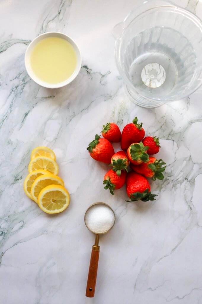 Fresh strawberries, sliced lemons, sugar, and lemon juice arranged beside a blender for homemade patriotic strawberry lemonade preparation.