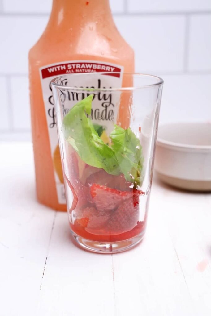 Fresh basil leaves added to a glass with sliced strawberries beside a bottle of Simply drink for homemade drink preparation.