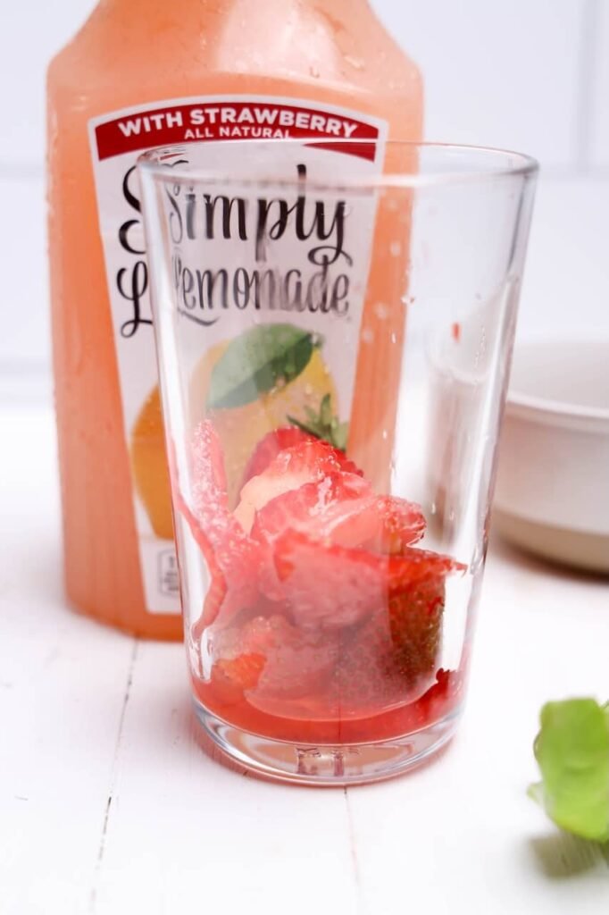 Glass filled with sugared sliced strawberries beside a bottle of Simply drink during preparation of homemade mocktail.