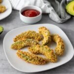 Golden brown Air Fryer Avocado Fries arranged on a white plate and served with a dipping sauce. The avocado wedges are coated in a crispy breadcrumb crust and cooked until crunchy on the outside while remaining creamy and tender inside, ready to serve as an appetizer or snack.