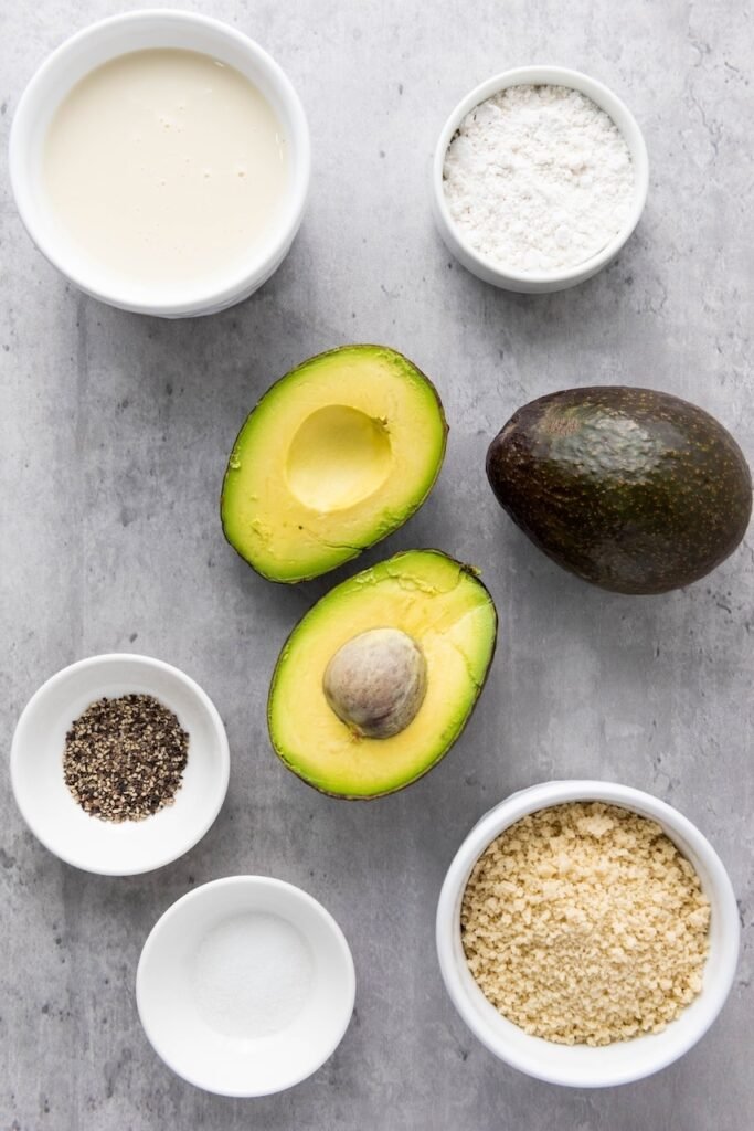 Ingredients for Air Fryer Avocado Fries arranged on a gray surface, including ripe avocados, soy milk, flour, panko breadcrumbs, salt, and black pepper before preparation.