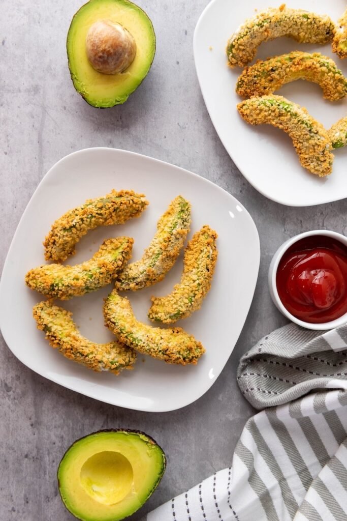 Overhead view of crispy Air Fryer Avocado Fries arranged on a white plate with a side of dipping sauce and fresh avocado halves. The breaded avocado wedges are golden brown and crunchy on the outside while remaining creamy inside, ready to serve as a vegan appetizer or snack.