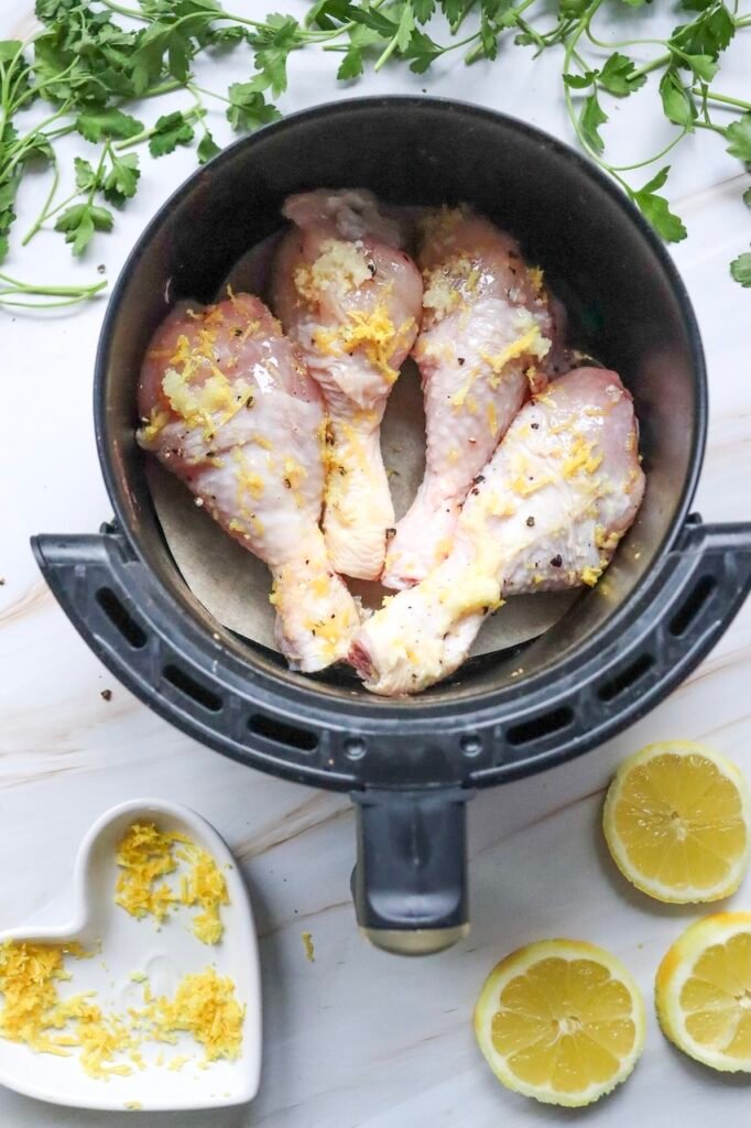 Marinated drumsticks arranged in a single layer inside a basket with lemon zest and garlic visible on the poultry. Fresh parsley, lemon halves, and extra lemon zest surround the basket on a white marble surface. This image shows the drumsticks prepared and ready to be air fried.