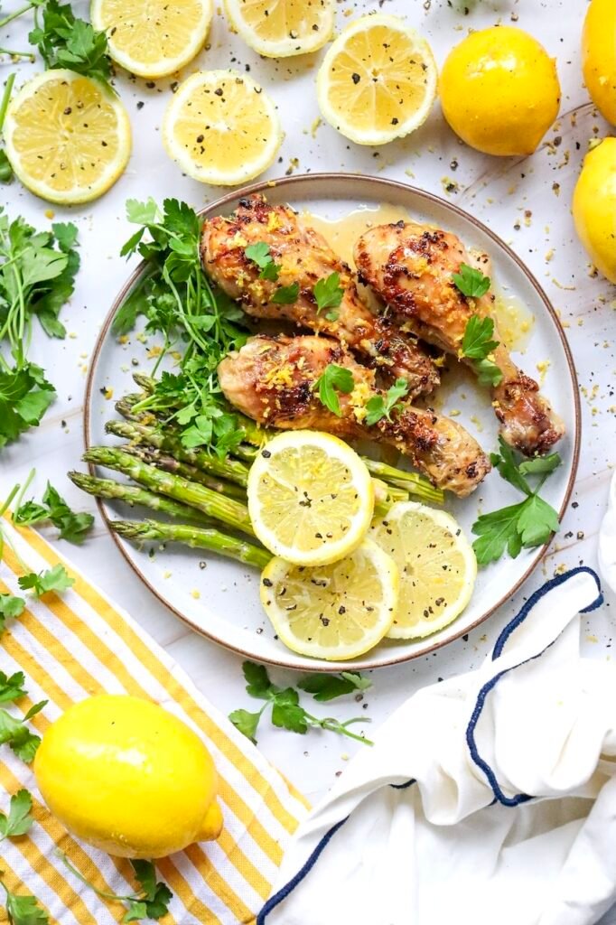 Overhead view of lemon herb air fryer chicken legs served with asparagus, fresh parsley, and sliced lemons on a plate. The chicken is golden brown and garnished with lemon zest, while fresh herbs and lemons surround the dish on a light marble surface. This image showcases the finished chicken drumsticks recipe ready to serve.