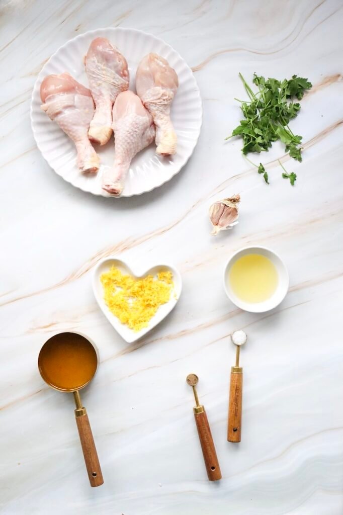Ingredients arranged on a white marble surface, including raw chicken drumsticks, fresh parsley, garlic, lemon zest, olive oil, chicken broth, salt, and pepper. This image shows the ingredients needed to prepare the marinade before seasoning.