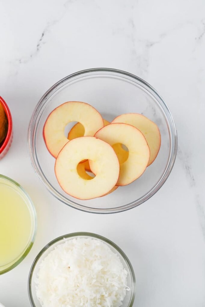 Overhead view of cored apple slices in a glass bowl during preparation for apple slice cookies, with bowls of lime juice and sweetened coconut flakes nearby on a white marble surface.