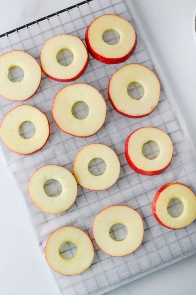 Overhead view of cored apple slices drying on a wire rack after soaking in lime juice, preparing them for peanut butter and toppings in an apple slice cookies recipe.