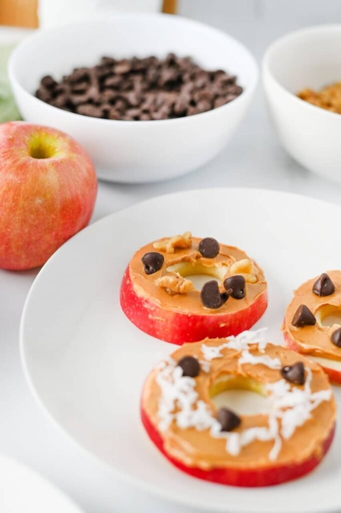 Close-up overhead view of apple slice cookies topped with peanut butter, chocolate chips, coconut flakes, and walnuts on a white plate, ready to serve as a healthy snack.