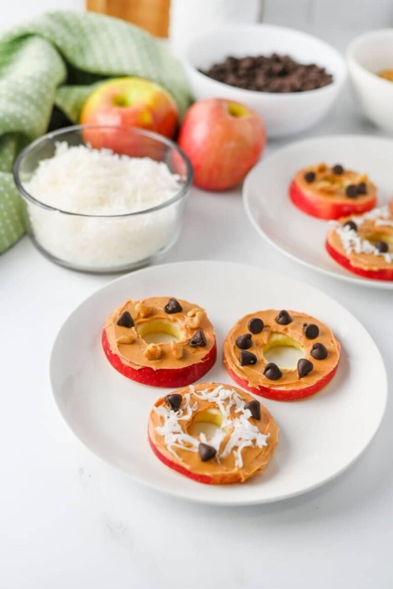 Three apple slice cookies topped with peanut butter, chocolate chips, coconut flakes, and walnuts on a white plate, with additional ingredients and apple slice cookies visible in the background.