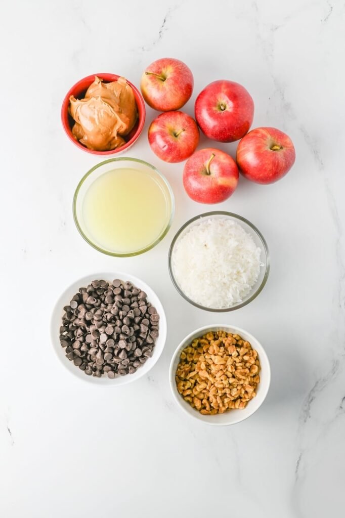 Overhead view of ingredients for apple slice cookies arranged on a white marble surface, including red apples, peanut butter, lime juice, chocolate chips, sweetened coconut flakes, and chopped walnuts before assembly.