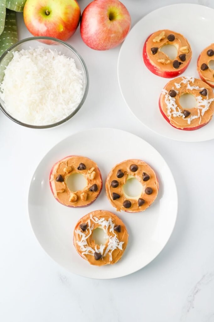 Overhead view of apple slice cookies topped with peanut butter, chocolate chips, coconut flakes, and walnuts on white plates, with fresh apples and ingredients arranged nearby.