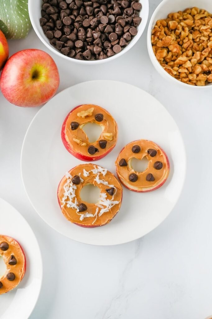 Overhead view of apple slice cookies topped with peanut butter, chocolate chips, coconut flakes, and walnuts on a white plate, with fresh apples and bowls of toppings nearby.