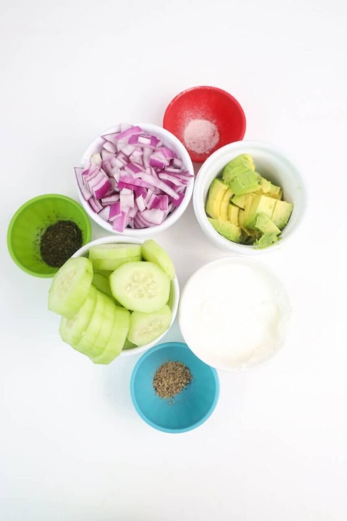 Overhead photo of the ingredients for avocado cucumber salad arranged in small bowls on a white background, including sliced cucumber, diced red onion, cubed avocado, sour cream, dill, salt, and black pepper before mixing.