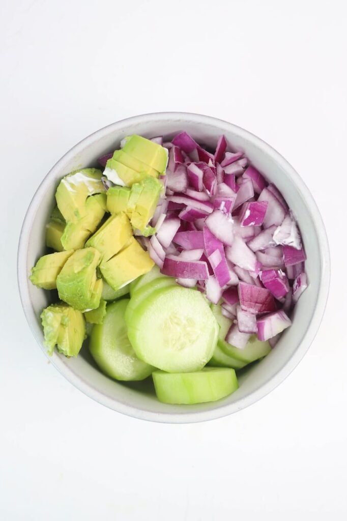 Overhead photo of sliced cucumbers, cubed avocado, and diced red onion combined in a white bowl as the first mixing step for avocado cucumber salad.