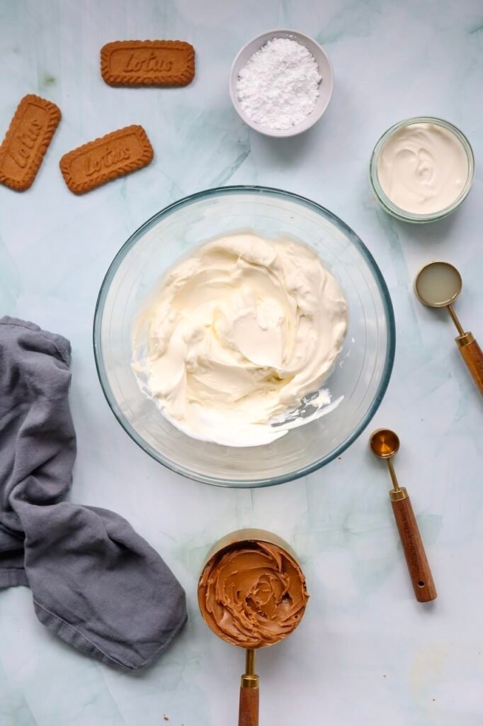 Overhead view of softened cream cheese beaten until smooth in a glass mixing bowl, surrounded by  cookies, spread, powdered sugar, whipping cream, vanilla extract, and lemon juice while preparing the filling for a no-bake dessert.