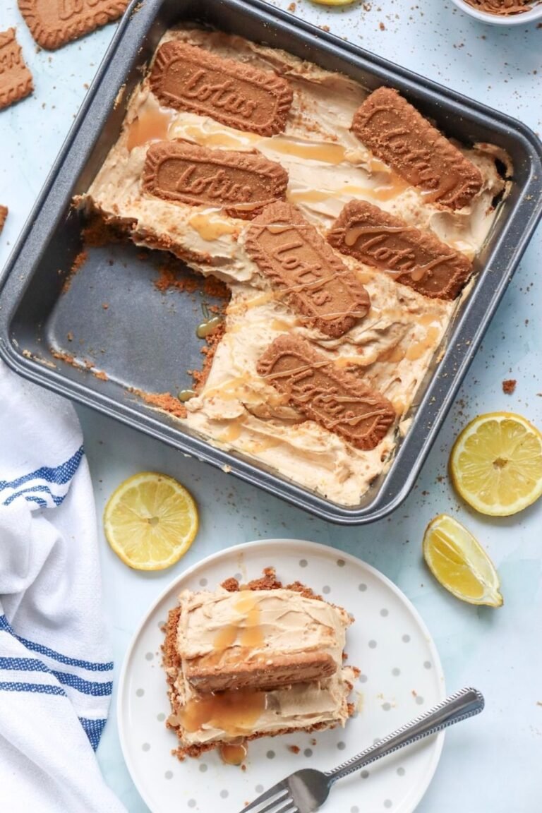 Overhead view of no-bake Biscoff cheesecake bars in a square baking pan with one serving removed to reveal the creamy cookie butter cheesecake filling and buttery Biscoff cookie crust. The cheesecake is topped with Lotus Biscoff cookies and a drizzle of melted Biscoff spread, with a plated cheesecake bar served alongside.