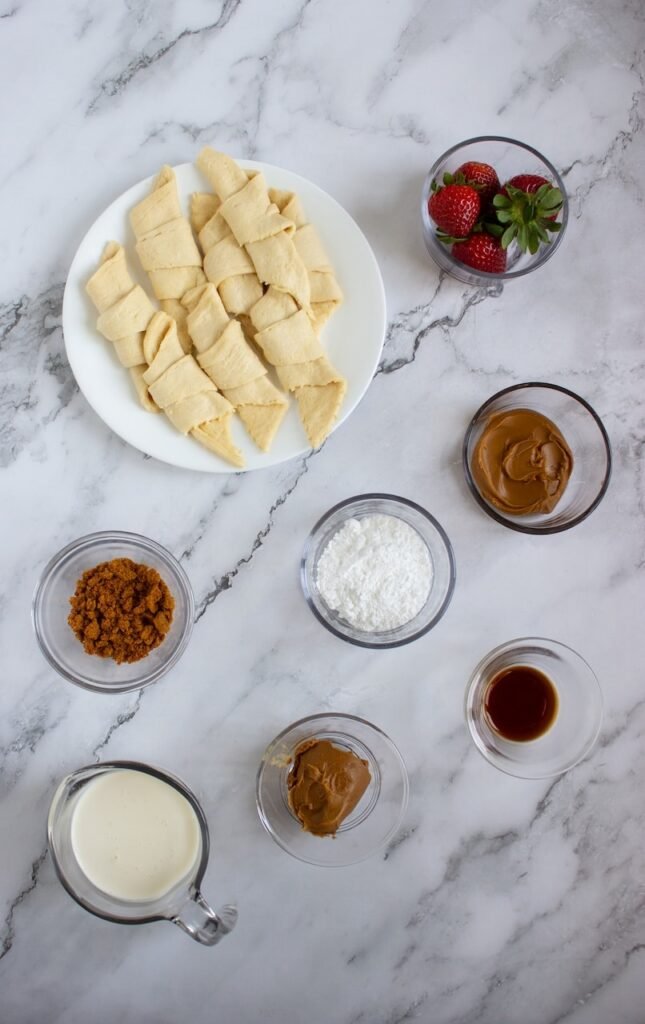 Ingredients for Biscoff strawberry croffles arranged on a marble surface, including crescent roll dough, fresh strawberries, Biscoff spread, heavy cream, powdered sugar, vanilla extract, and crushed Biscoff cookies.