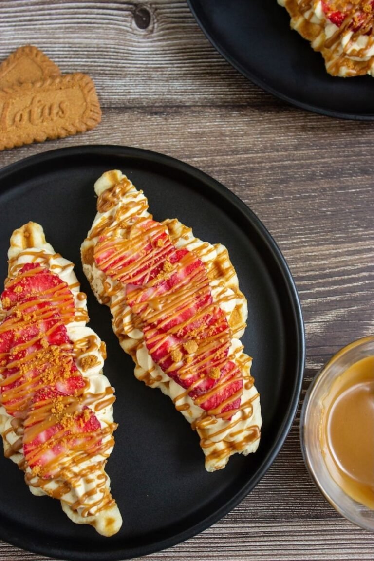 Two finished Biscoff strawberry croffles served on black plates and topped with piped Biscoff whipped cream, fresh sliced strawberries, melted Biscoff drizzle, and crushed Biscoff cookies. The croffles have a crispy golden exterior with creamy, fruity, and cookie butter toppings, ready to serve as an easy brunch or dessert.