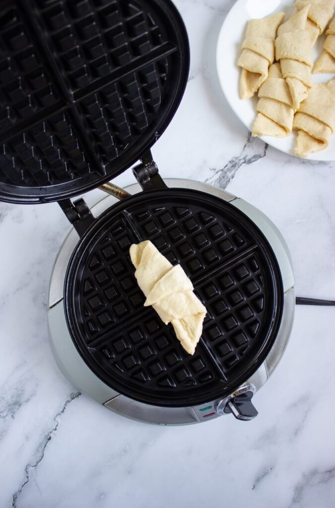 A rolled crescent dough triangle placed inside an open waffle iron on a marble countertop, showing the process of cooking homemade breakfast. This step helps create the croffles’ crispy golden exterior while keeping the inside soft, buttery, and flaky.