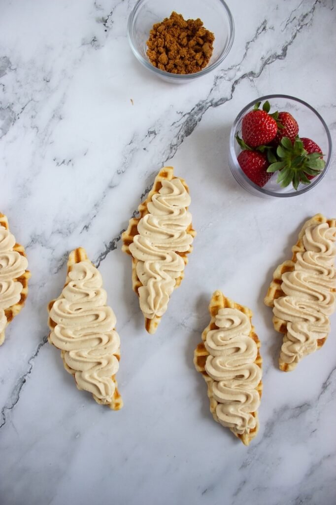 Freshly cooked croffles arranged on a marble surface and topped with piped Biscoff whipped cream, with bowls of fresh strawberries and crushed Biscoff cookies nearby for finishing. This step shows the assembly process before adding the final toppings for Biscoff strawberry croffles.