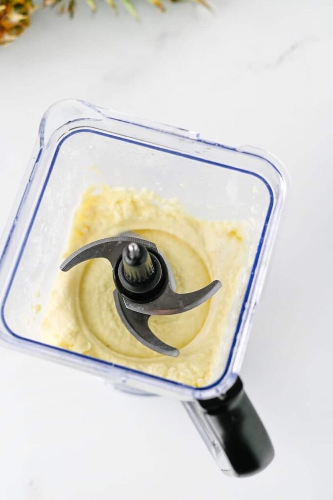 Smooth and creamy soft serve mixture blended in a blender container, showing the thick soft-serve texture made from frozen pineapple, pineapple juice, vanilla ice cream, and lemon juice before being piped into serving cups.