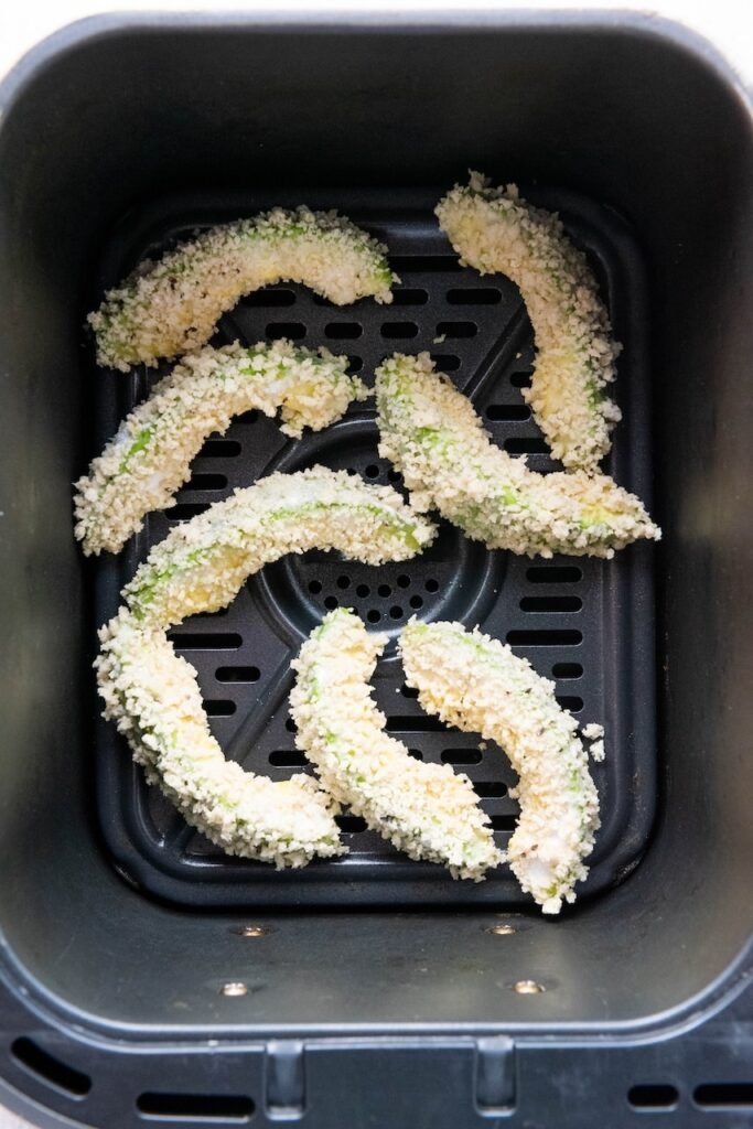 Breaded avocado wedges arranged in a single layer inside an air fryer basket before cooking. Leaving space between each avocado slice allows hot air to circulate evenly, helping the panko coating become golden brown and crispy while the avocado stays creamy inside.